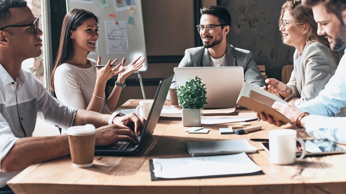 Junges Gründerteam diskutiert lachend am Tisch mit Laptops und Whiteboard, Kundengewinnung beginnt im direkten Gespräch